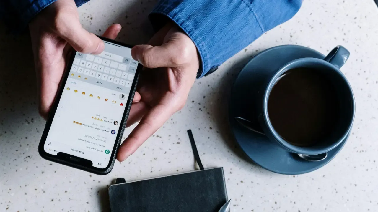 En la foto: Persona con un celular en mano, en la mesa una libreta y una taza de café